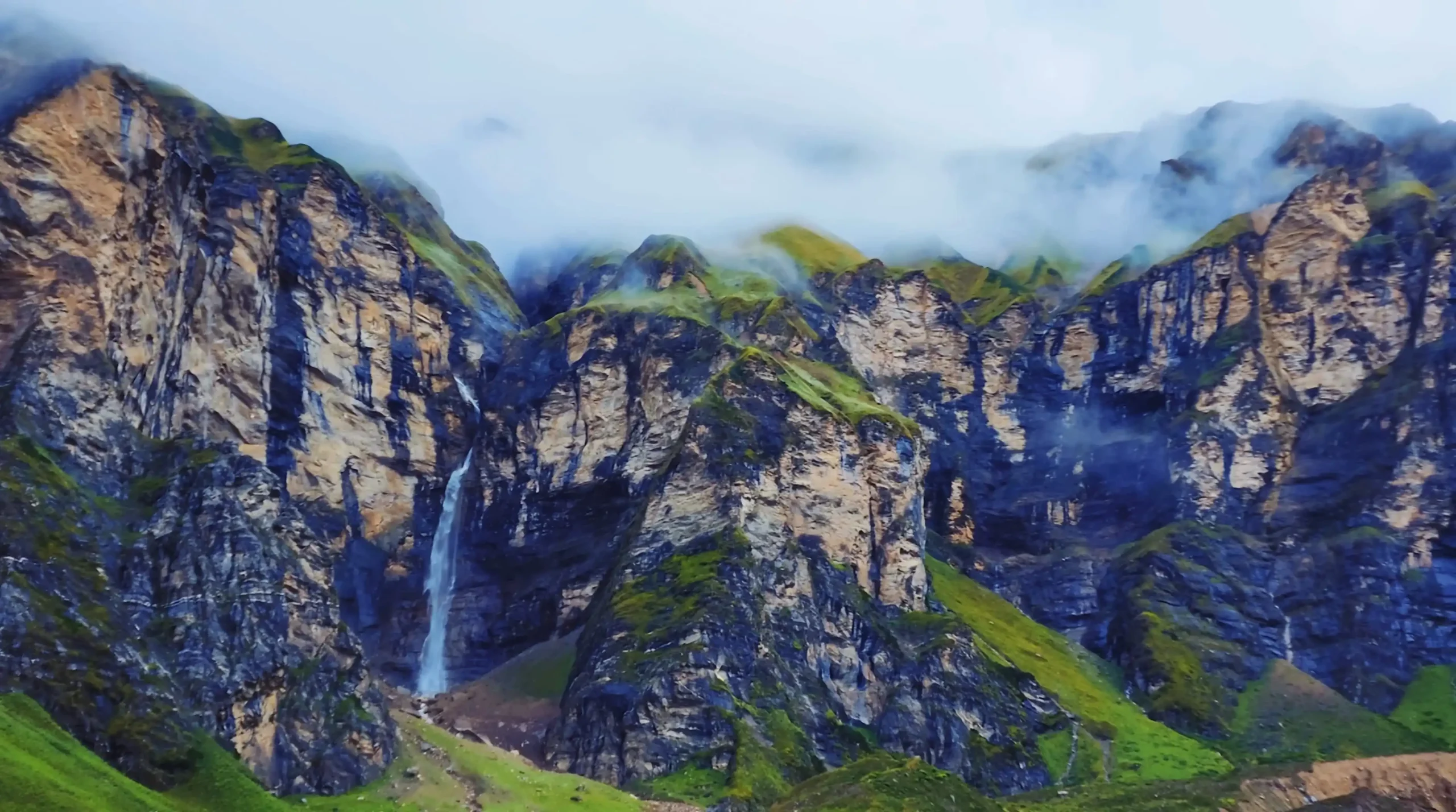 saipal base camp: A lush green mountainside with a narrow waterfall cascading down exposed rock, set against towering snow-capped peaks in the background.